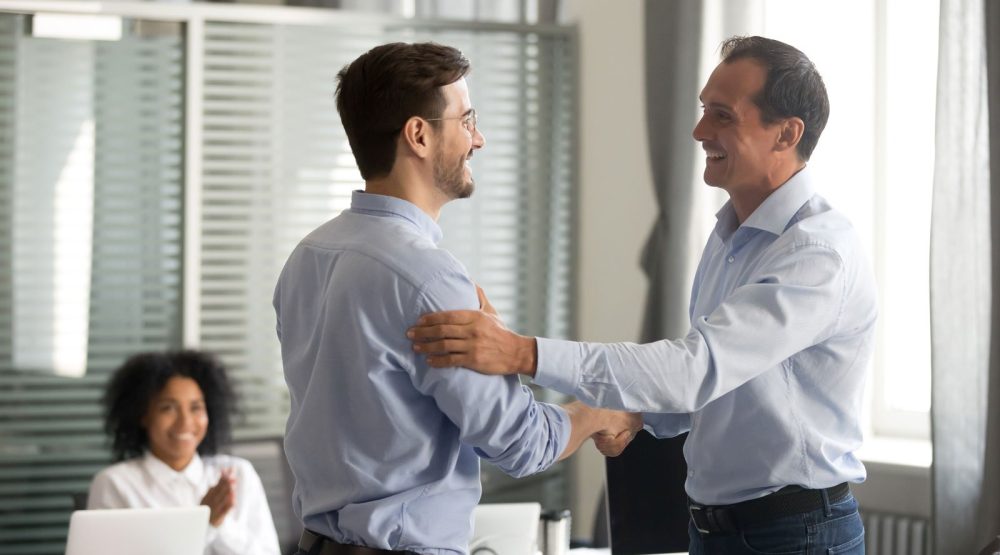 Two colleagues shaking hands while another colleague applauds in an office.