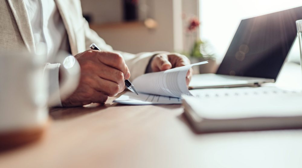 Person reviewing and signing paperwork beside a laptop.